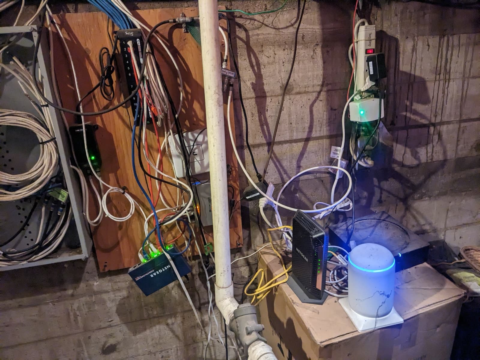 Tangled network wiring on a basement plywood board, with a modem and mesh router sitting on a cardboard box